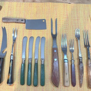 May include: An assortment of vintage cutlery and serving utensils, including knives, forks, and a bar spoon. The collection features various handle designs, including wood, green, and dark brown. A cleaver is also present.