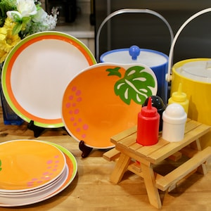 May include: A set of orange and green patterned plates with a small wooden picnic table holding condiment bottles. The table has a yellow and blue cooler behind it.