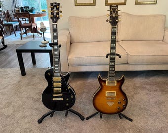 May include: Two electric guitars standing on black floor stands. The left guitar is a black Gibson with gold hardware and a white-bound fretboard. The right guitar is a sunburst-finish Ibanez with gold hardware and a double-cutaway body shape.