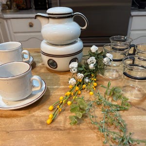 May include: A white ceramic teapot with a matching warmer, two cups and saucers, and four glass mugs with black handles and rims. The set is arranged on a wooden surface with decorative floral accents.