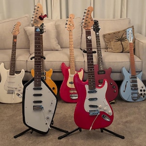 May include: A collection of seven electric guitars standing on floor stands. The guitars feature various body shapes and colors, including a unique white-bodied guitar with black trim, a bright pink guitar, a light blue hollow-body, and others.
