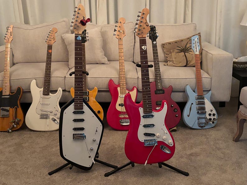 May include: A collection of seven electric guitars standing on floor stands. The guitars feature various body shapes and colors, including a unique white-bodied guitar with black trim, a bright pink guitar, a light blue hollow-body, and others.