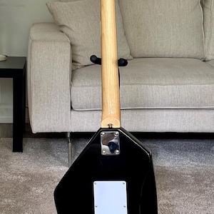 May include: The back of a black electric guitar with a unique angular body shape. It features a light-colored wooden neck, a chrome neck plate, and a white rectangular back plate with six string holes. The headstock displays the Vox logo.