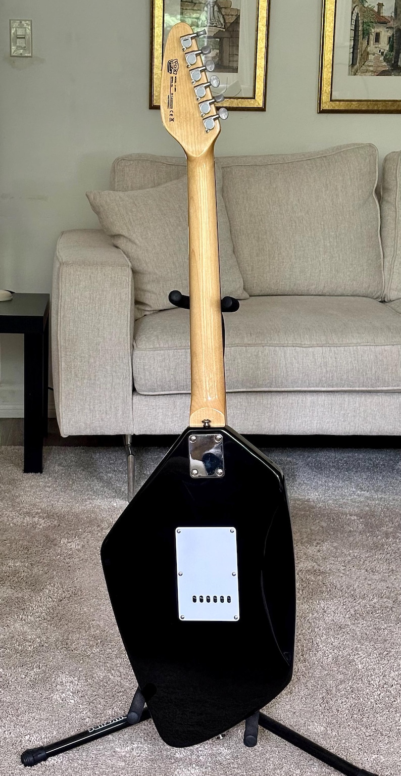 May include: The back of a black electric guitar with a unique angular body shape. It features a light-colored wooden neck, a chrome neck plate, and a white rectangular back plate with six string holes. The headstock displays the Vox logo.