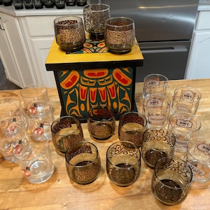 May include: A set of 14 clear glass tumblers with various designs. The tumblers are arranged on a wooden box with a colorful design. The box has a carved design of a Native American totem pole.