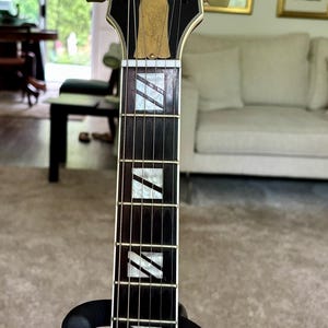 May include: A Gibson Les Paul Supreme electric guitar headstock and neck featuring a black finish, gold-plated tuning pegs, a mother-of-pearl inlay logo, and a truss rod cover engraved with Les Paul. The dark fretboard has white block inlays.