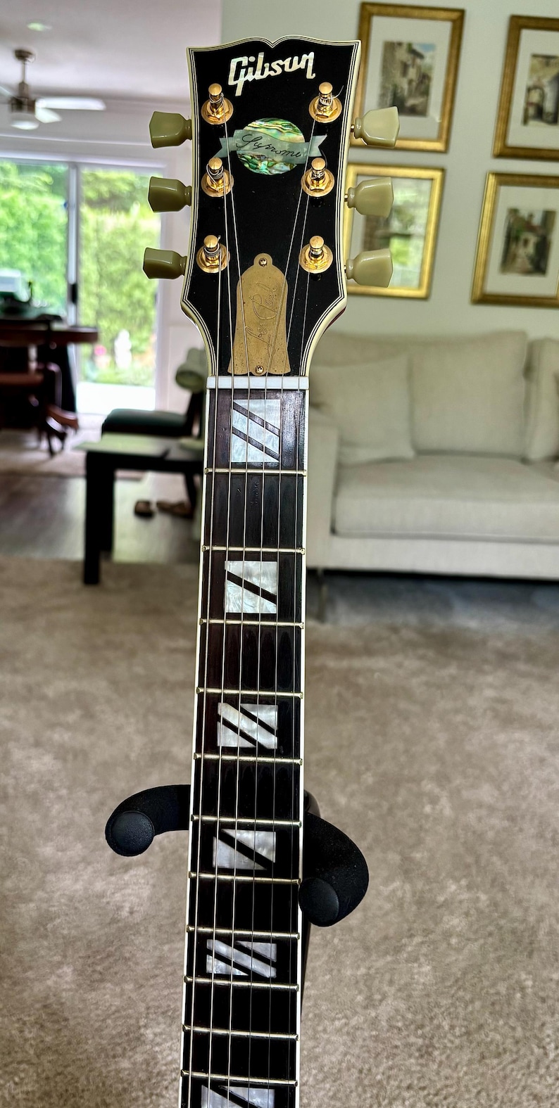 May include: A Gibson Les Paul Supreme electric guitar headstock and neck featuring a black finish, gold-plated tuning pegs, a mother-of-pearl inlay logo, and a truss rod cover engraved with Les Paul. The dark fretboard has white block inlays.