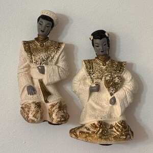 Puede incluir: Dos placas de pared de cerámica que representan figuras con vestimenta tradicional. Cada figura tiene rostro y manos grises, vistiendo túnicas de color crema con detalles texturizados pintados en oro. Una figura lleva una gorra y la otra un accesorio floral.