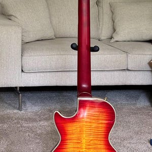 May include: An electric guitar shown from the back, featuring a cherry sunburst finish with a visible wood grain pattern. The neck is dark red, and the headstock includes six gold-toned tuning pegs with cream-colored buttons and a serial number.
