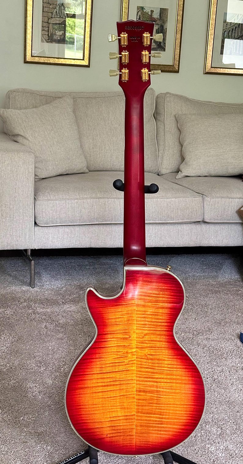 May include: An electric guitar shown from the back, featuring a cherry sunburst finish with a visible wood grain pattern. The neck is dark red, and the headstock includes six gold-toned tuning pegs with cream-colored buttons and a serial number.