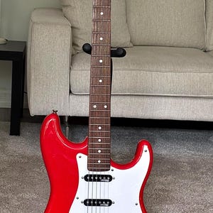 May include: A bright red electric guitar with a white pickguard, a dark wood fretboard with white dot inlays, and a light wood headstock featuring the brand name Eko. The instrument includes three black single-coil pickups and chrome hardware.