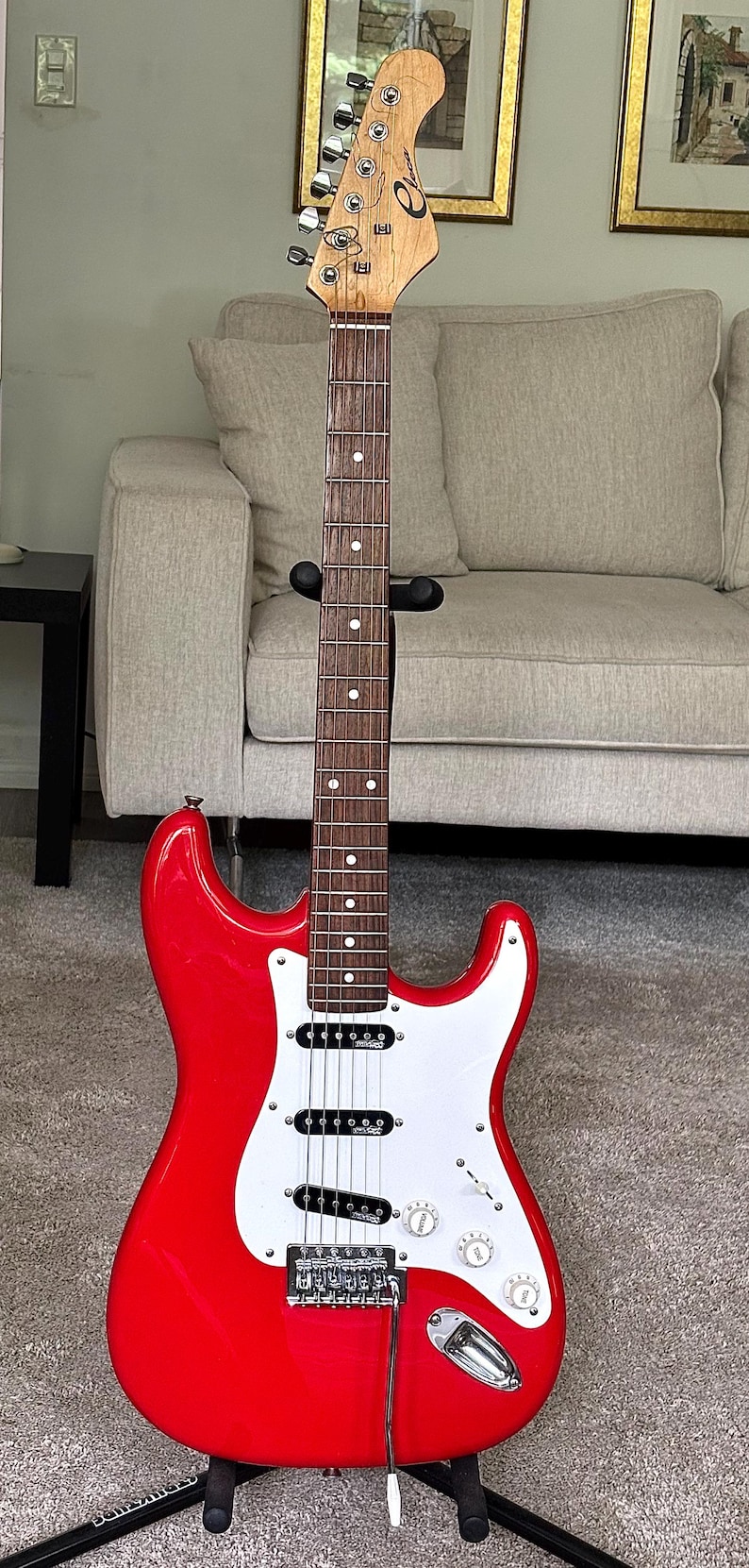 May include: A bright red electric guitar with a white pickguard, a dark wood fretboard with white dot inlays, and a light wood headstock featuring the brand name Eko. The instrument includes three black single-coil pickups and chrome hardware.