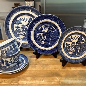 Puede incluir: Una colección de porcelana azul y blanca con diseño Willow, que incluye platos de varios tamaños y una taza de té con platillo. Los platos se exhiben sobre soportes negros. La porcelana presenta un diseño de paisaje detallado.