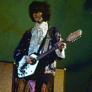 May include: A musician with dark, voluminous hair wears a long, dark-colored coat featuring a floral pattern and a white ruffled shirt underneath. The musician plays a white, double-neck electric guitar with a patterned strap against a green background.