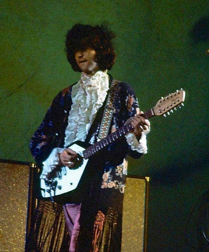 May include: A musician with dark, voluminous hair wears a long, dark-colored coat featuring a floral pattern and a white ruffled shirt underneath. The musician plays a white, double-neck electric guitar with a patterned strap against a green background.