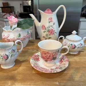 Puede incluir: Un juego de té vintage con tetera, jarra de crema, azucarero, taza de té y platillo. El juego es blanco con estampados florales de rosas rosas y ribetes dorados. También hay un jarrón con una flor rosa.
