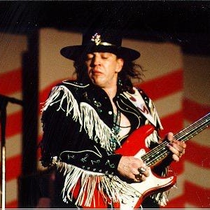 May include: A musician wearing a black hat with a decorative pin and a black jacket featuring long white fringe on the sleeves and shoulders. The musician is playing a bright red electric guitar with a white pickguard and a light-colored neck.