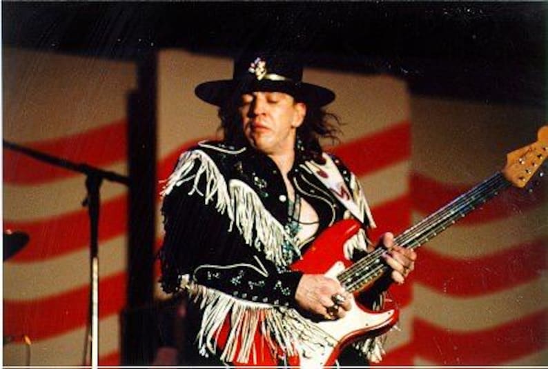 May include: A musician wearing a black hat with a decorative pin and a black jacket featuring long white fringe on the sleeves and shoulders. The musician is playing a bright red electric guitar with a white pickguard and a light-colored neck.