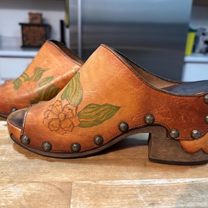 May include: A pair of open-toe clogs in a warm, light brown leather. The clogs feature a floral design with green leaves and a flower. The sides are adorned with metal studs, and the heels are approximately 3 inches high.