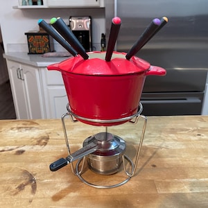 May include: Red cast iron fondue pot with a stainless steel stand and six fondue forks with colorful handles. The pot is on a wooden table.