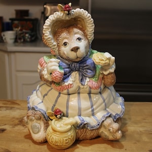 Puede incluir: Una jarra de galletas de cerámica con forma de oso que lleva un vestido azul y blanco con un lazo rojo. El oso sostiene una galleta y una cesta de fruta. La jarra tiene una olla de miel en la parte inferior.