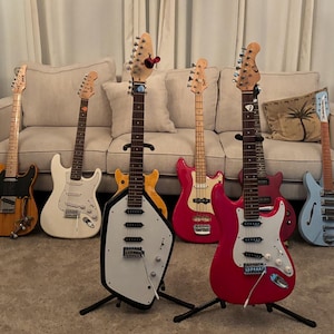 May include: A collection of seven electric guitars and basses in various colors and shapes, including a natural wood finish, white, yellow, red, pink, and light blue models, arranged in a row with some on stands and others leaning against a sofa.