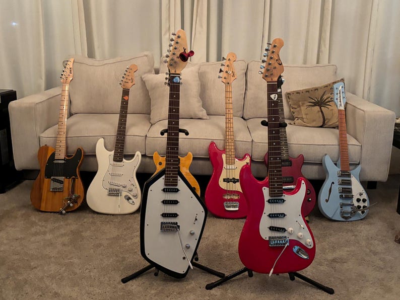 May include: A collection of seven electric guitars and basses in various colors and shapes, including a natural wood finish, white, yellow, red, pink, and light blue models, arranged in a row with some on stands and others leaning against a sofa.
