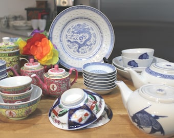 Op de afbeelding: Een verzameling keramisch serviesgoed, waaronder een grote blauw-witte drakenbord, twee theepotten, diverse kleine kommen met blauwe vismotieven en kleurrijke kommen met gele, rode en groene patronen en decoratieve randen.