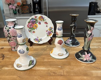 May include: A collection of ceramic home decor items including a floral vase, a scalloped plate with pansy motifs, and four candlestick holders. Two holders are white with seashell patterns, one is black, and one is black with floral designs.