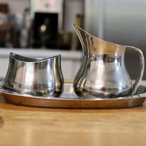 May include: A silver creamer and sugar bowl set on a silver tray. The creamer and sugar bowl are both round and have a smooth, shiny finish.