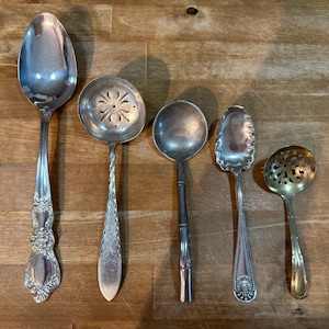 Könnte beinhalten: Eine Sammlung von fünf antiken Servierlöffeln aus Silber, jedes mit einem einzigartigen Design. Die Löffel variieren in Größe und Form, darunter ein Schöpflöffel und eine Schöpfkelle. Sie sind auf einer Holzoberfläche angeordnet.