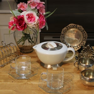 Puede incluir: Una tetera blanca con tapa y base plateadas, dos tazas de té de vidrio transparente con platillos a juego, dos jarras de crema plateadas, un tostadora de plata y un plato decorativo plateado con un diseño floral. La tetera está sobre una mesa de madera con un ramo de flores rosas en un jarrón de vidrio.