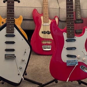 May include: A collection of electric guitars and a bass guitar featuring a variety of body shapes and colors, including a black and white shield-shaped guitar, a bright red double-cutaway guitar, a red bass, and a yellow wood-grain guitar.