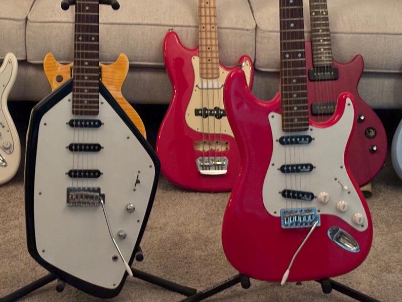 May include: A collection of electric guitars and a bass guitar featuring a variety of body shapes and colors, including a black and white shield-shaped guitar, a bright red double-cutaway guitar, a red bass, and a yellow wood-grain guitar.