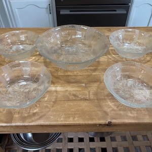 May include: Five clear glass bowls of different sizes are displayed on a wooden surface. The bowls feature a textured, ribbed exterior and a floral pattern inside. The largest bowl is centrally positioned, with four smaller bowls around it.