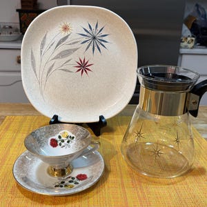 May include: A vintage tea set featuring a cream-coloured, square plate with floral and starburst designs. A clear glass carafe with gold accents and a black handle is next to a teacup and saucer with floral details.