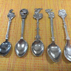 Peut inclure: Cinq cuillères décoratives en argent avec des motifs détaillés sur les manches. Les cuillères présentent divers éléments décoratifs, notamment des animaux et des motifs floraux. Elles sont disposées sur une surface texturée jaune-orangé.