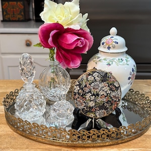 Pode incluir: Uma bandeja dourada com um design floral decorativo, incluindo um espelho redondo, dois frascos de perfume de vidro transparente e um espelho compacto com padrão floral. A bandeja é decorada com um design floral e tem uma borda festonada. Os frascos de perfume são de vidro transparente e têm uma superfície texturizada. O espelho compacto é redondo e tem um design floral. A bandeja está sobre uma superfície de madeira.