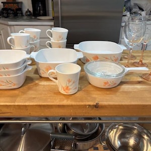 May include: A selection of white ceramic cookware and drinkware featuring a floral pattern. Includes casserole dishes, mugs, and a saucepan with a glass lid. Wine glasses and champagne flutes are also present on a wooden table.