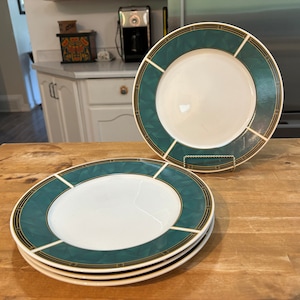 May include: Three green and gold rimmed dinner plates stacked on a wooden table. The plates have a white centre and a green and gold rim.