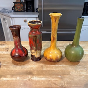 Könnte beinhalten: Eine Sammlung von vier Keramikvasen, die in einer Reihe angeordnet sind. Die erste ist eine kleine bauchige Vase in Dunkelrot und Braun, die zweite eine höhere zylindrische Vase in Rot und Beige, die dritte eine hohe gelb-braune Vase und die vierte ist grün.