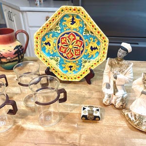 May include: A collection of decorative items on a wooden table. Includes a painted pitcher, a colorful ceramic plate, glass mugs with brown handles, two Asian-style figurines, and a small decorative box.