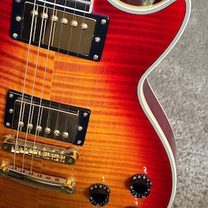 May include: An electric guitar with a cherry sunburst finish featuring a flamed maple top, gold-colored hardware, two humbucker pickups, a tune-o-matic bridge, and four black control knobs with white numbering on the body of the instrument.