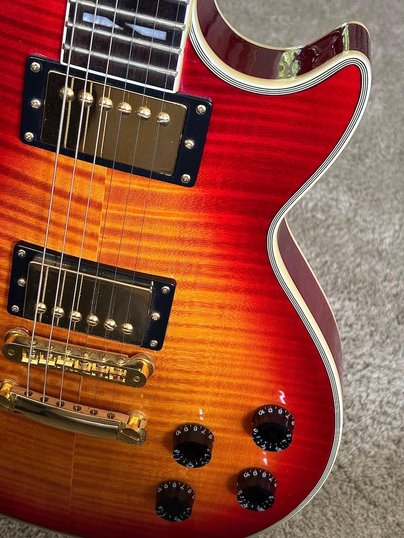 May include: An electric guitar with a cherry sunburst finish featuring a flamed maple top, gold-colored hardware, two humbucker pickups, a tune-o-matic bridge, and four black control knobs with white numbering on the body of the instrument.