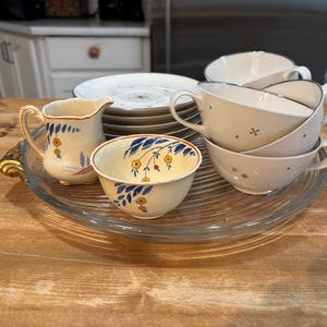 Puede incluir: Un conjunto de vajilla de cerámica que incluye una pequeña jarra, un cuenco y varias tazas con platos. La jarra y el cuenco presentan motivos de hojas azules y flores amarillas. Las tazas y platos son de color blanco roto con estrellas.