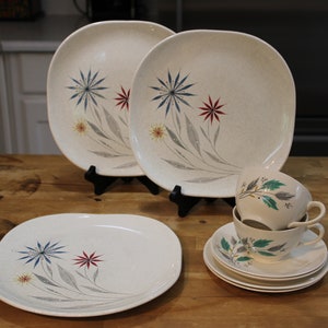 Può includere: Un set di piatti vintage color crema con un design floreale. Il set include piatti quadrati, tazze da tè e piattini. Il motivo floreale presenta esplosioni di stelle blu, rosse e gialle con foglie grigie. Le stoviglie sono disposte su una superficie di legno.