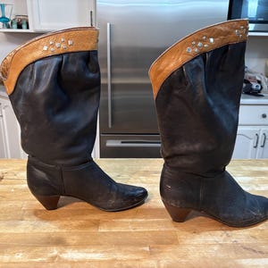 Puede incluir: Un par de botas de vaquero de cuero negro con la parte superior de cuero marrón y detalles de tachuelas plateadas. Las botas tienen un tacón de altura media y se exhiben sobre una superficie de madera. Estilo vintage.