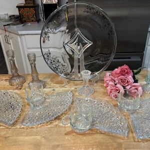 May include: A collection of vintage glassware and decorative items. Includes a large glass serving platter with silver accents, several candle holders, fan-shaped plates, and small drinking glasses. A bouquet of pink roses adds a touch of color.