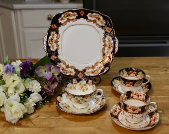 May include: A set of three vintage teacups and saucers with a blue and gold floral pattern. The cups and saucers are arranged on a wooden table with a bouquet of flowers in the foreground.