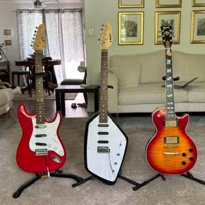 May include: Three electric guitars standing on individual black stands. The left is a red double-cutaway guitar with a white pickguard. The center is a white, angular, asymmetrical guitar. The right is a sunburst finish single-cutaway guitar.